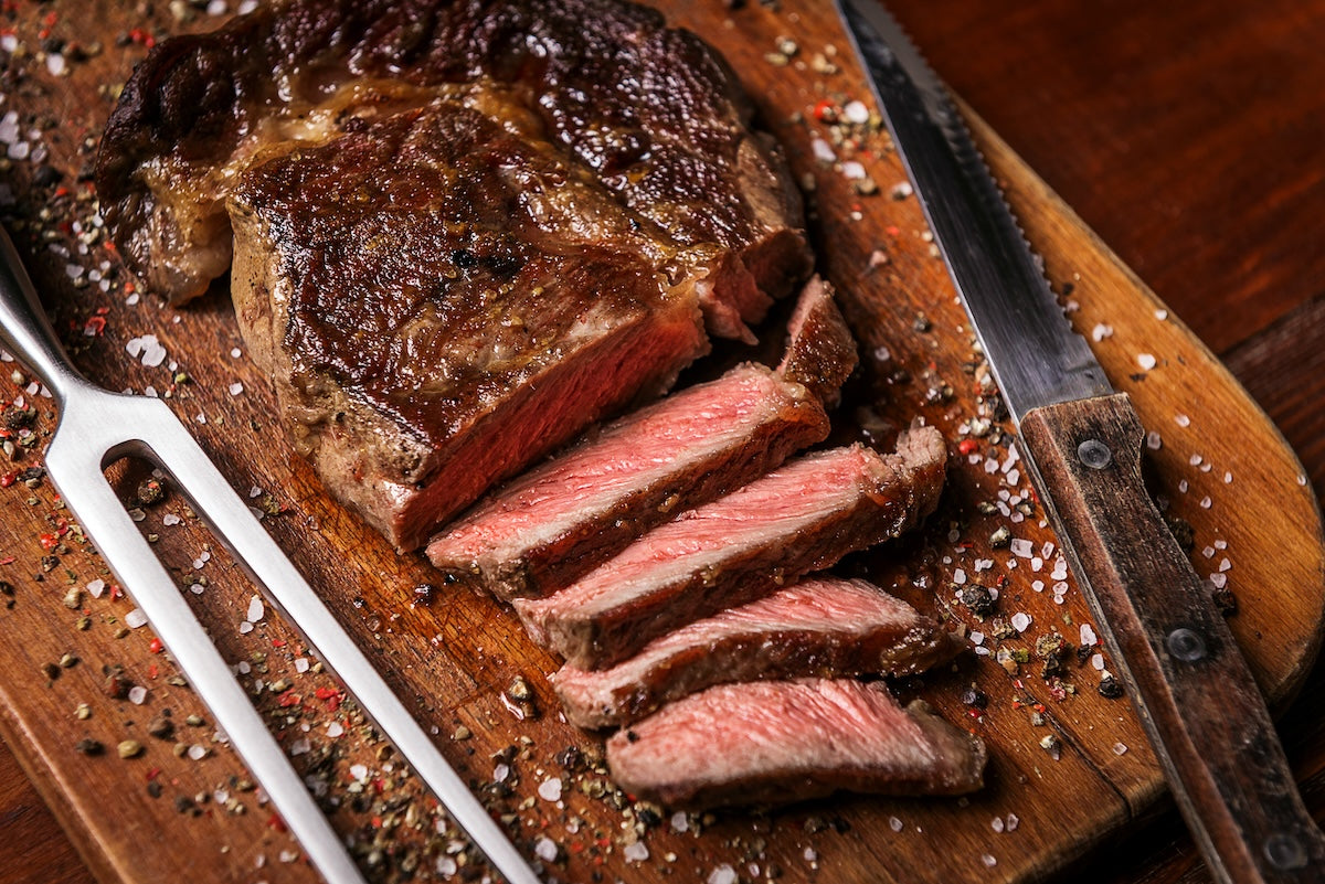 Sliced steak on a wooden cutting board with a knife and fork, surrounded by spices.